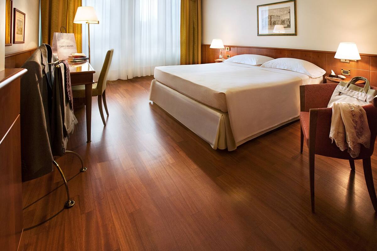 Hotellihuone, jossa on suuri sänky, kaksi yöpöytälamppua, kirjoituspöytä lampun kanssa, tuoli ja naulakko.
