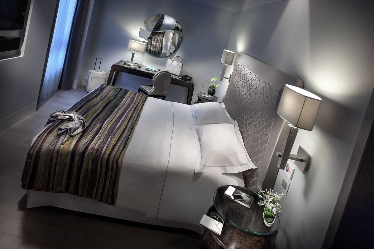 Moderni hotellihuone, jossa on siististi pedattu sänky, työpöytä tuolilla, yöpöytä lampun kanssa ja suuri peili seinällä.