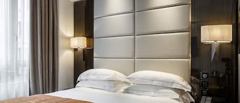 Una habitación de hotel moderna con una cama grande, dos lámparas de noche, un cabecero con panel de control y vistas de la ciudad a través de amplias ventanas.