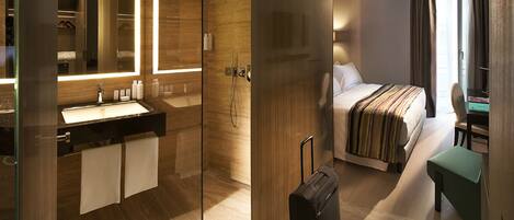 A modern hotel room with a glass shower enclosure, a large bed with a striped blanket, a desk with a chair, and a suitcase on the floor.