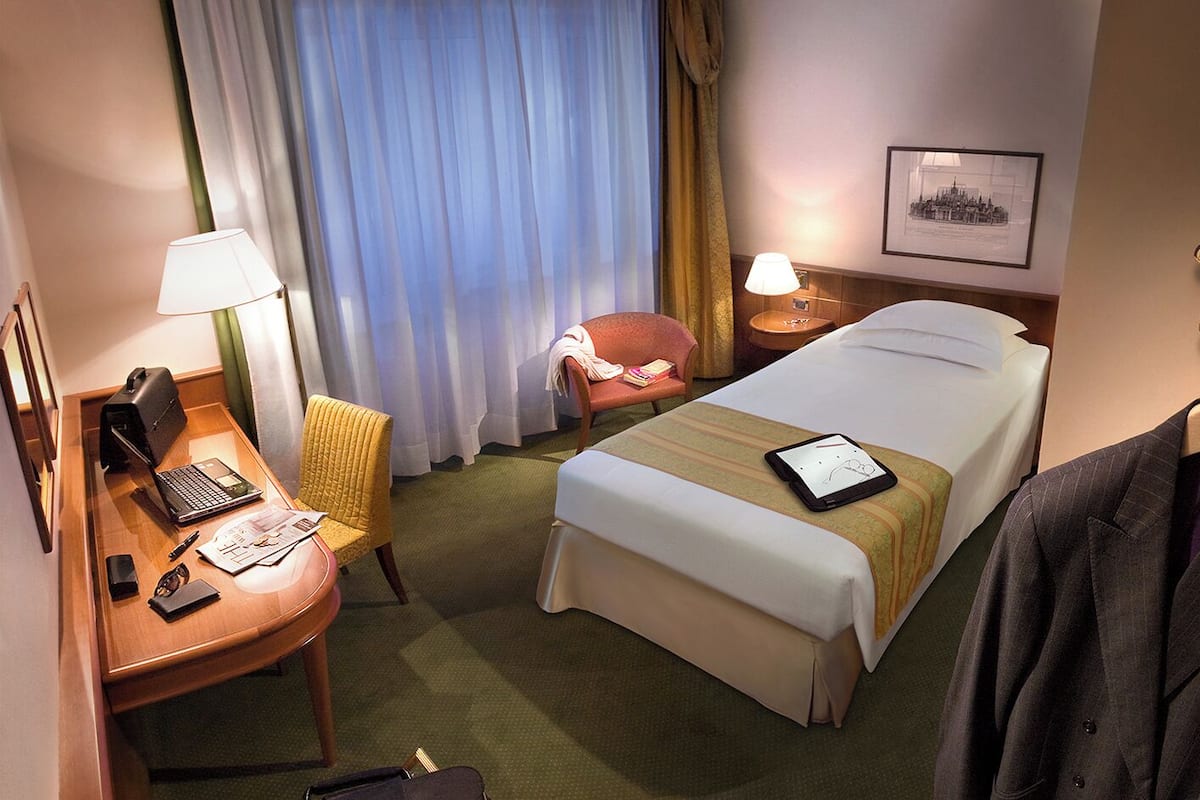 Hotellihuone, jossa on sänky, työpöytä kannettavalla tietokoneella, tuoli, yöpöytä lampun kanssa ja seinällä oleva kehystetty kuva.