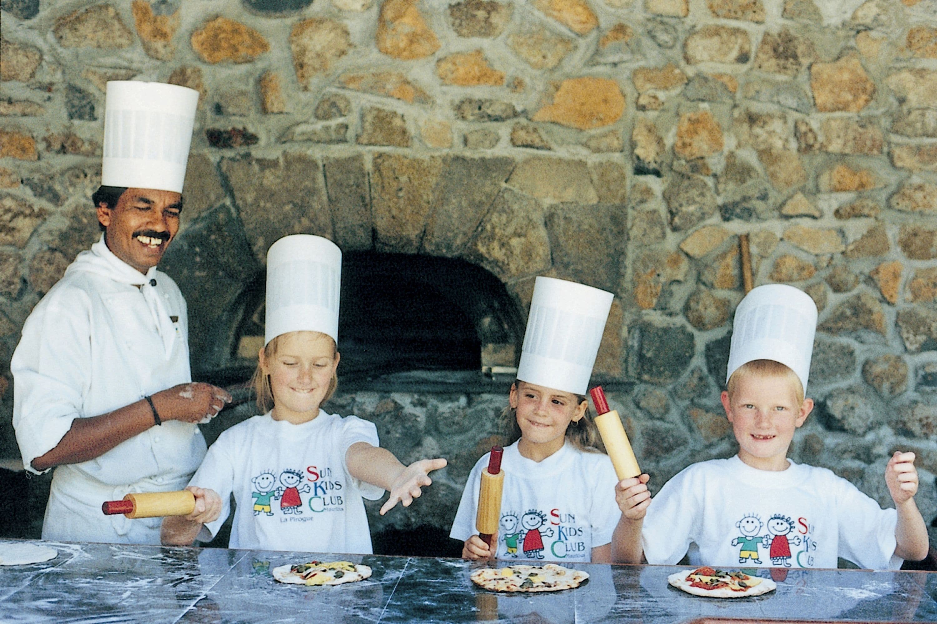 Un groupe de personnes portant des toques de chef, debout devant un mur de pierre avec un four à pizza.