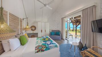 Une chambre avec un lit, un ventilateur de plafond, une télévision, un balcon avec vue sur la mer et un coin repas avec des chaises et une table.