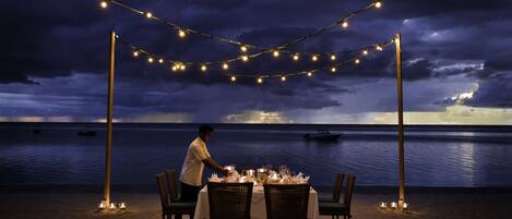 An outdoor dining setup with string lights, a table, chairs, and candles, overlooking a body of water.