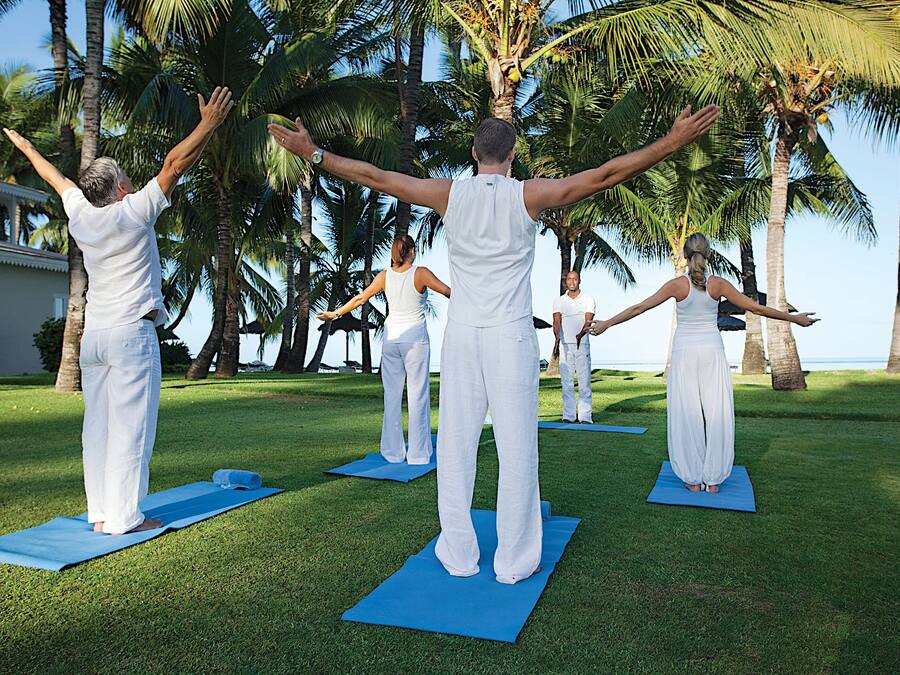 Un gruppo di persone che praticano yoga su tappetini blu in un'area erbosa, con palme e un edificio sullo sfondo.