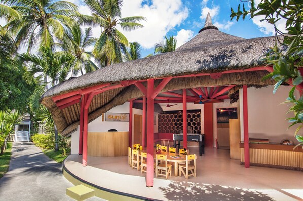 A thatched-roof pavilion with red pillars, a reception desk, and a children's play area with yellow chairs.