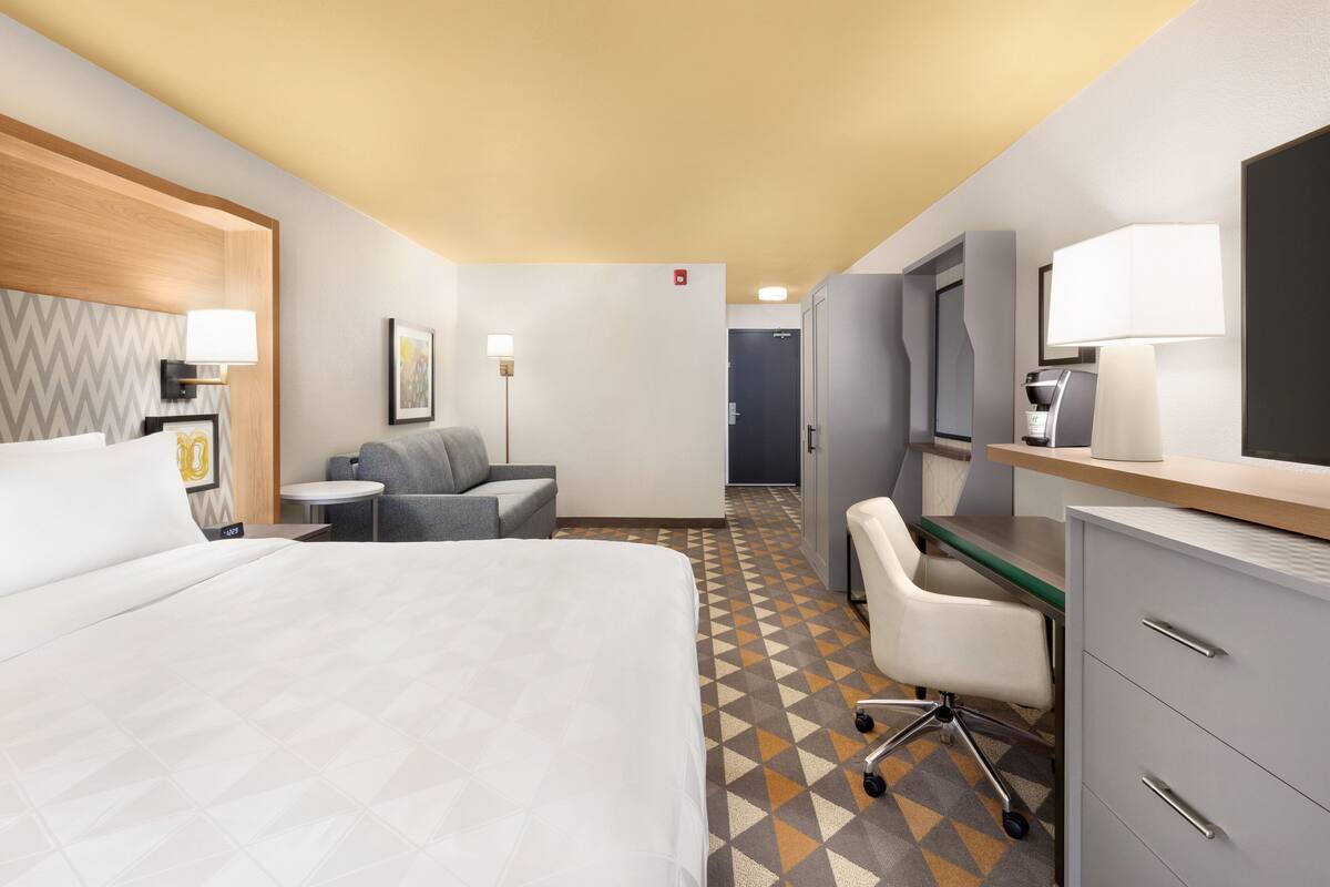 Ein Hotelzimmer mit Bett, Schreibtisch, Stuhl und einem kleinen Tisch. Auf dem Schreibtisch steht eine Lampe und an der Wand hängt ein Bild.