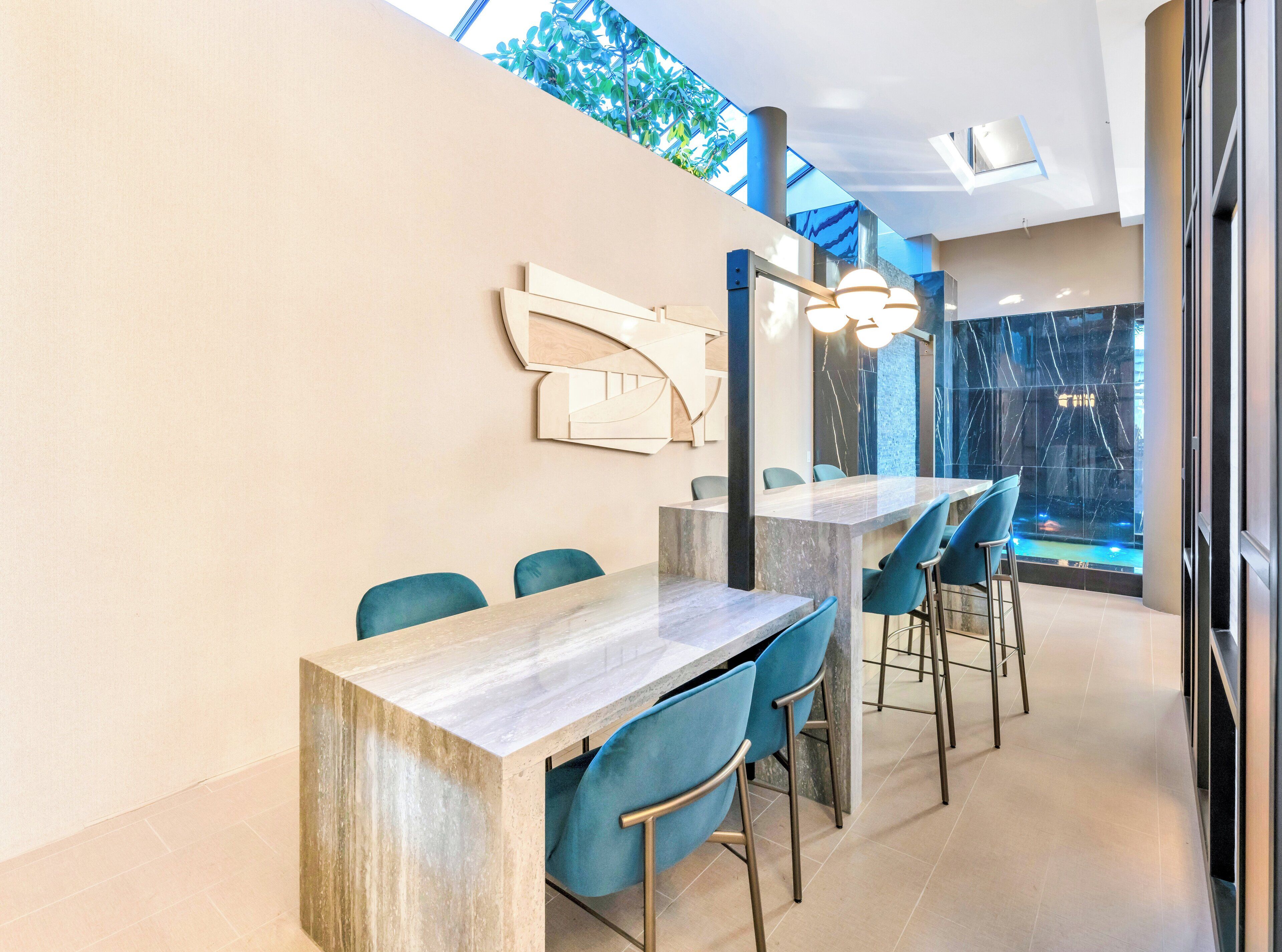 A modern dining area with a long table and blue chairs, a decorative wall piece, and a view of a waterfall through a glass wall.