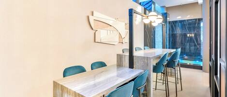 A modern dining area with a long table and blue chairs, a decorative wall piece, and a view of a waterfall through a glass wall.