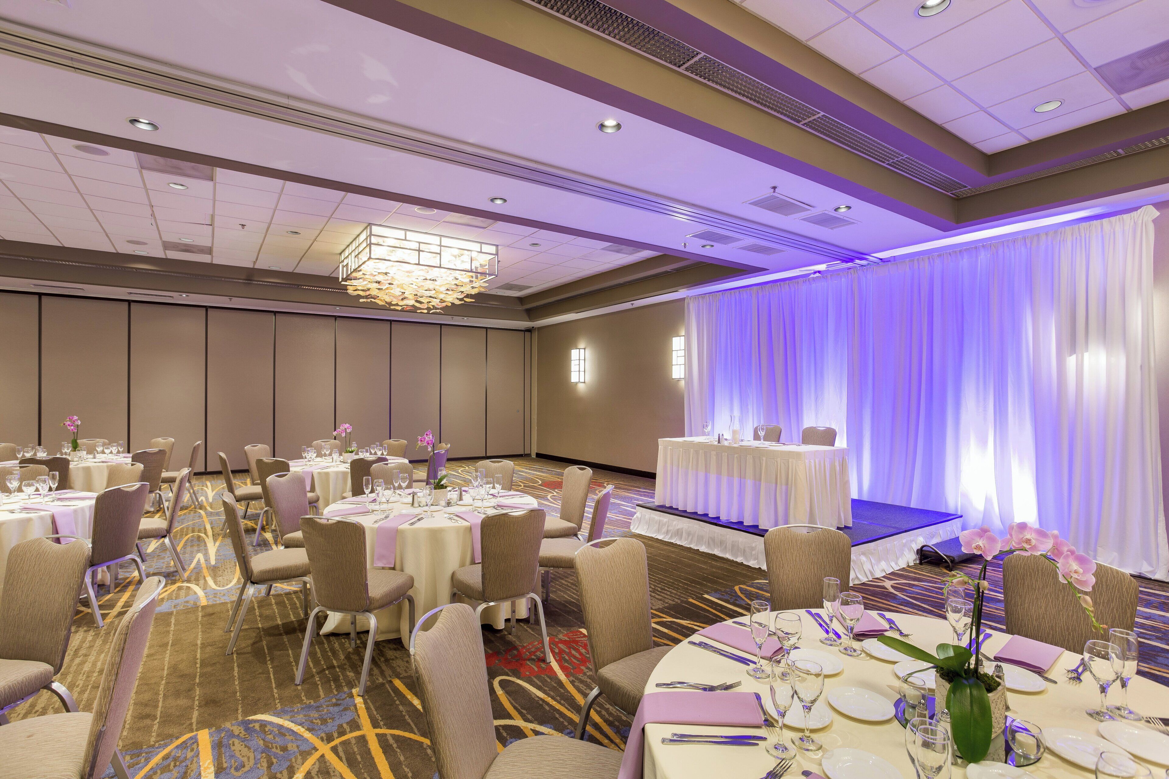 Un salón de banquetes con mesas redondas dispuestas para cenar, un escenario con cortinas y un candelabro.