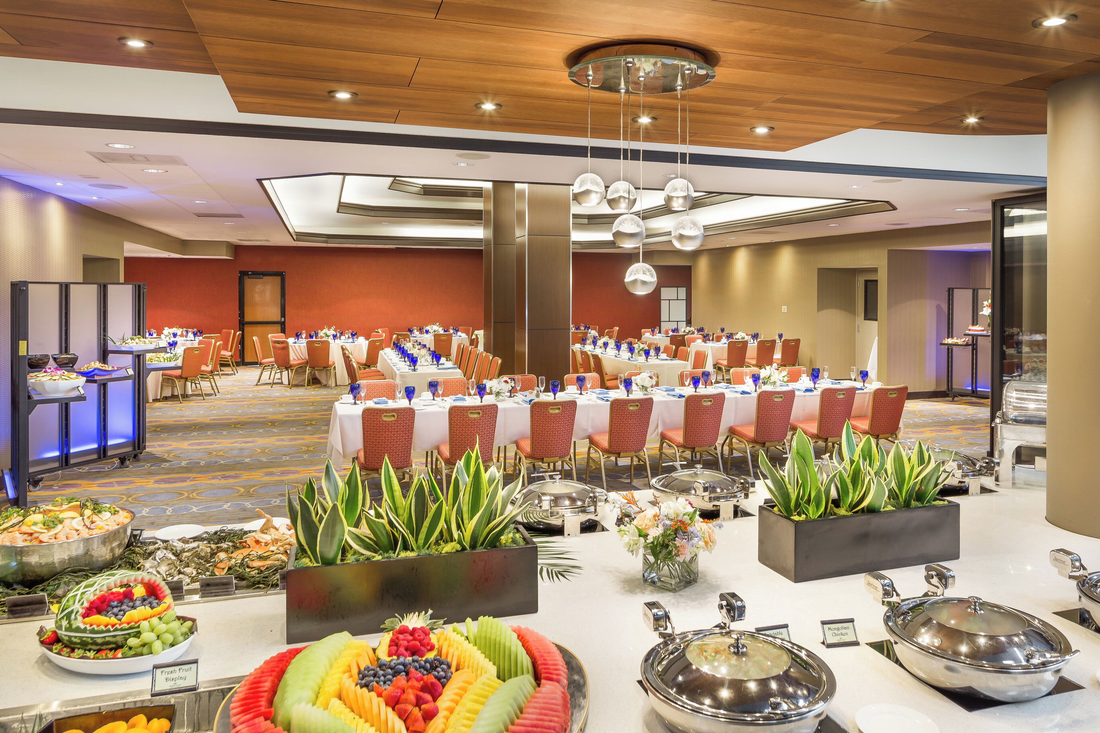 Salão de festas com mesas redondas preparadas para refeições, buffet com diversos pratos e teto com iluminação embutida.