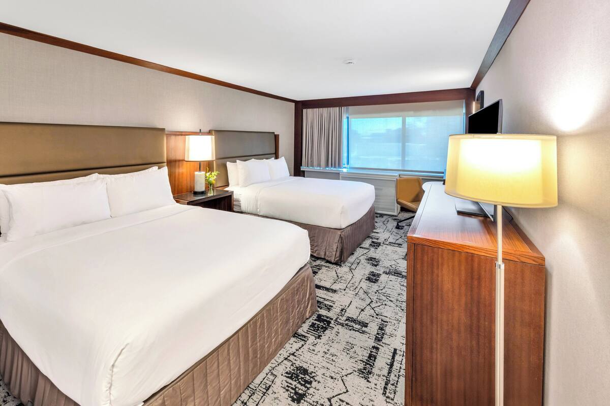 A hotel room with two beds, a desk, a TV, and a window with curtains.