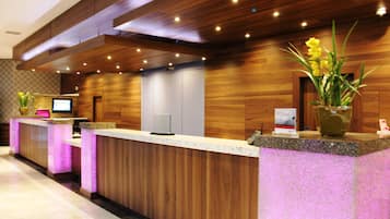 A hotel reception area with a wooden reception desk, a digital screen, and a potted plant.