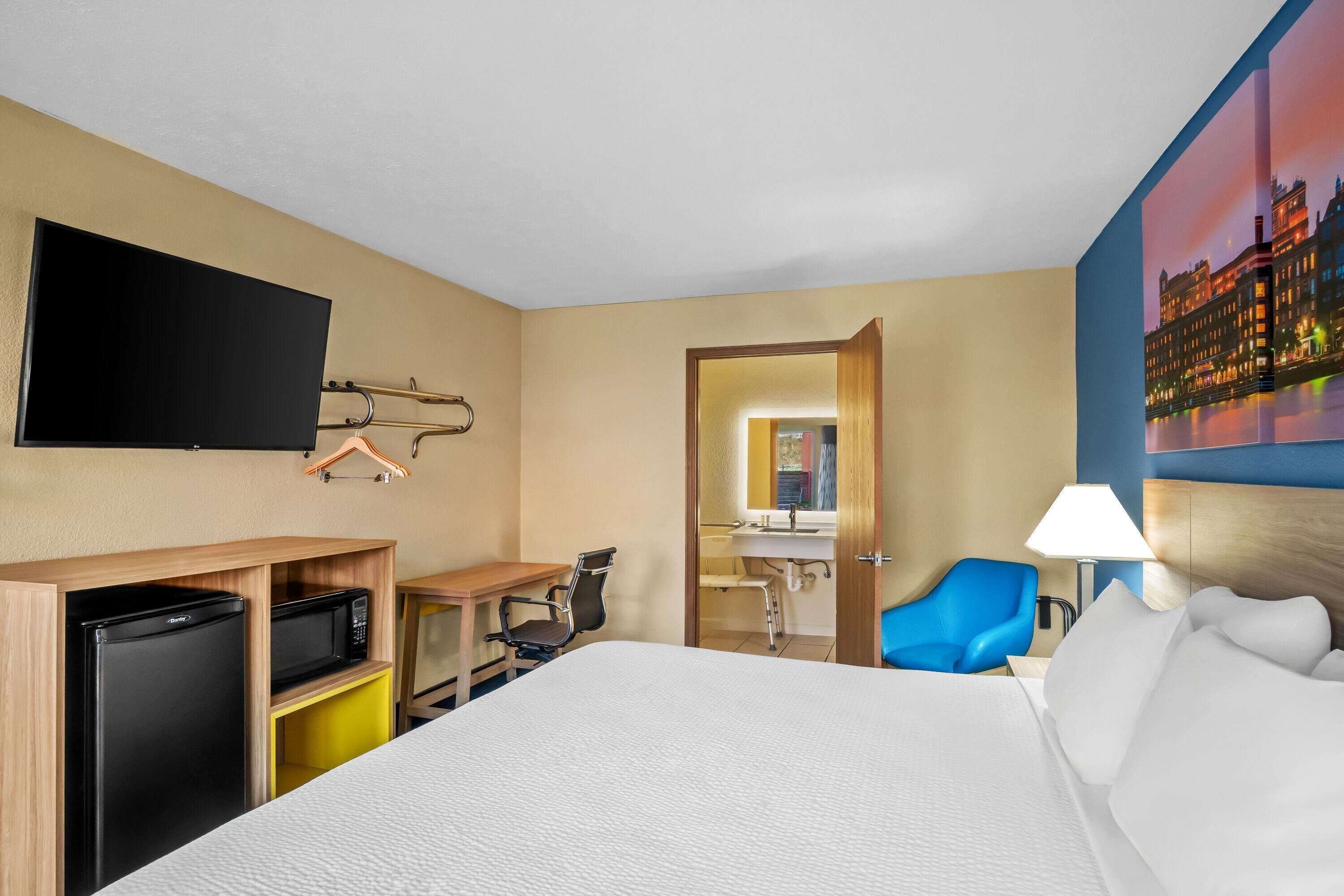 Habitación de hotel con cama, escritorio con computadora, televisor, refrigerador, silla, baño y un cuadro en la pared.