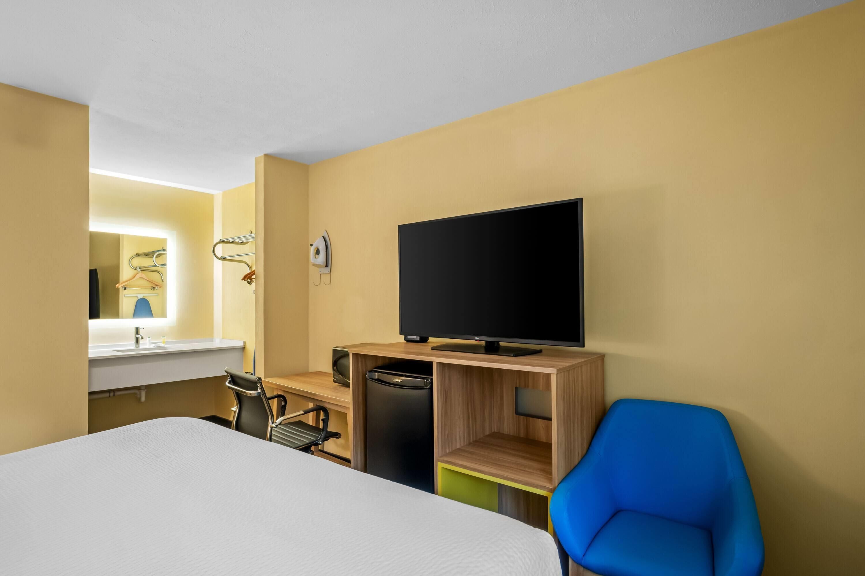 Habitación de hotel con cama, escritorio con televisor, silla, espejo y estante pequeño.