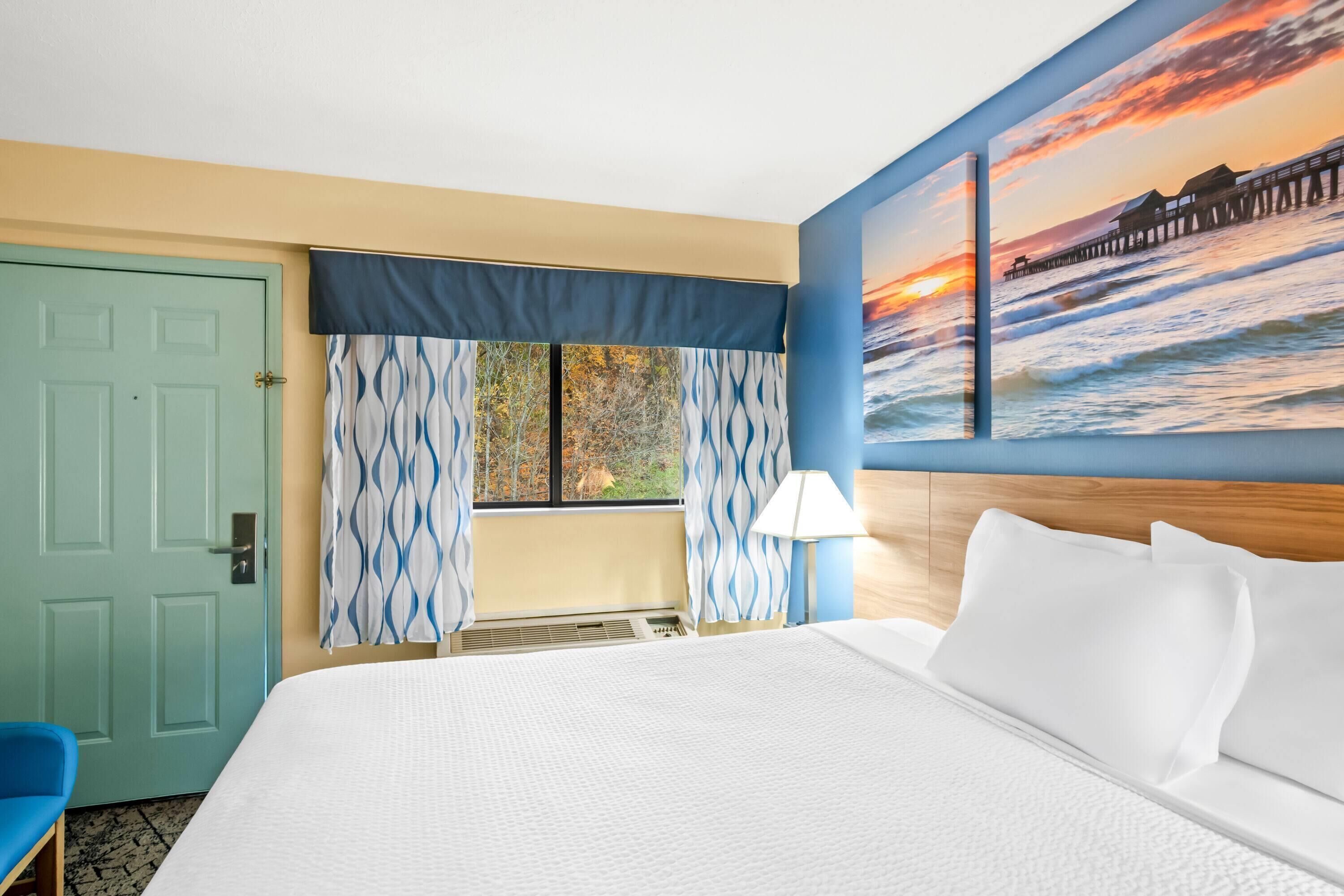 A hotel room with a bed, a window with curtains, a painting of a sunset over the ocean, and a blue door.