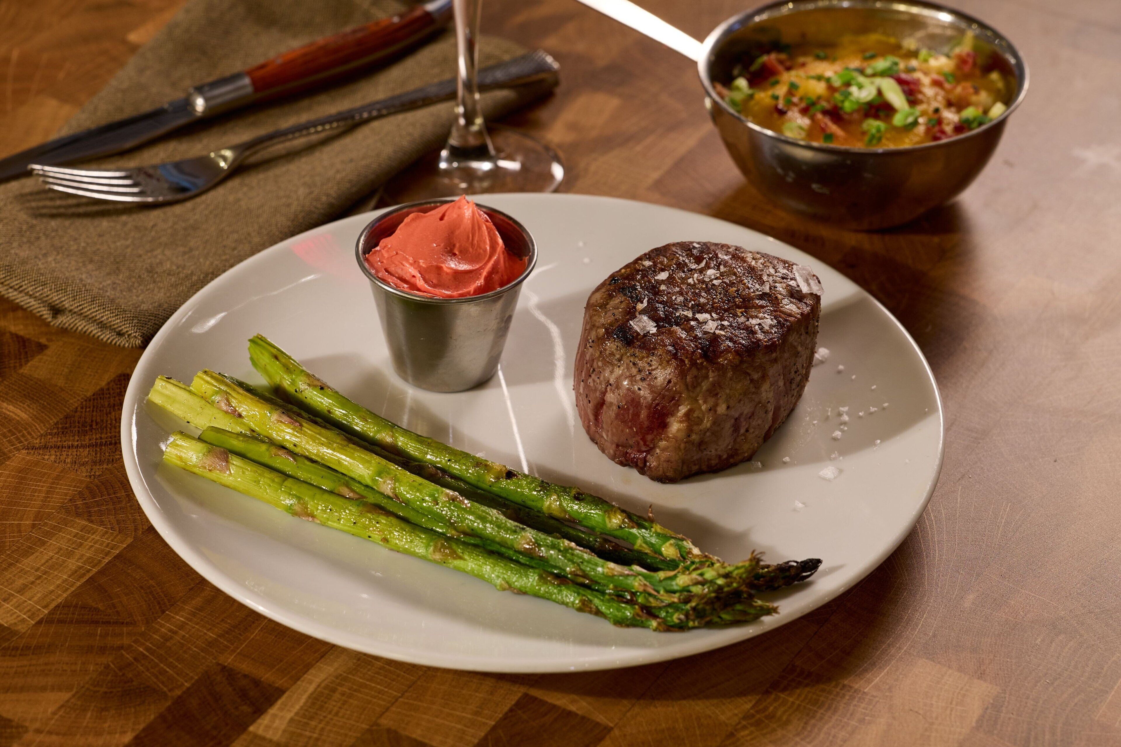 A plate with a steak, asparagus, and a small cup of sauce on a wooden table.