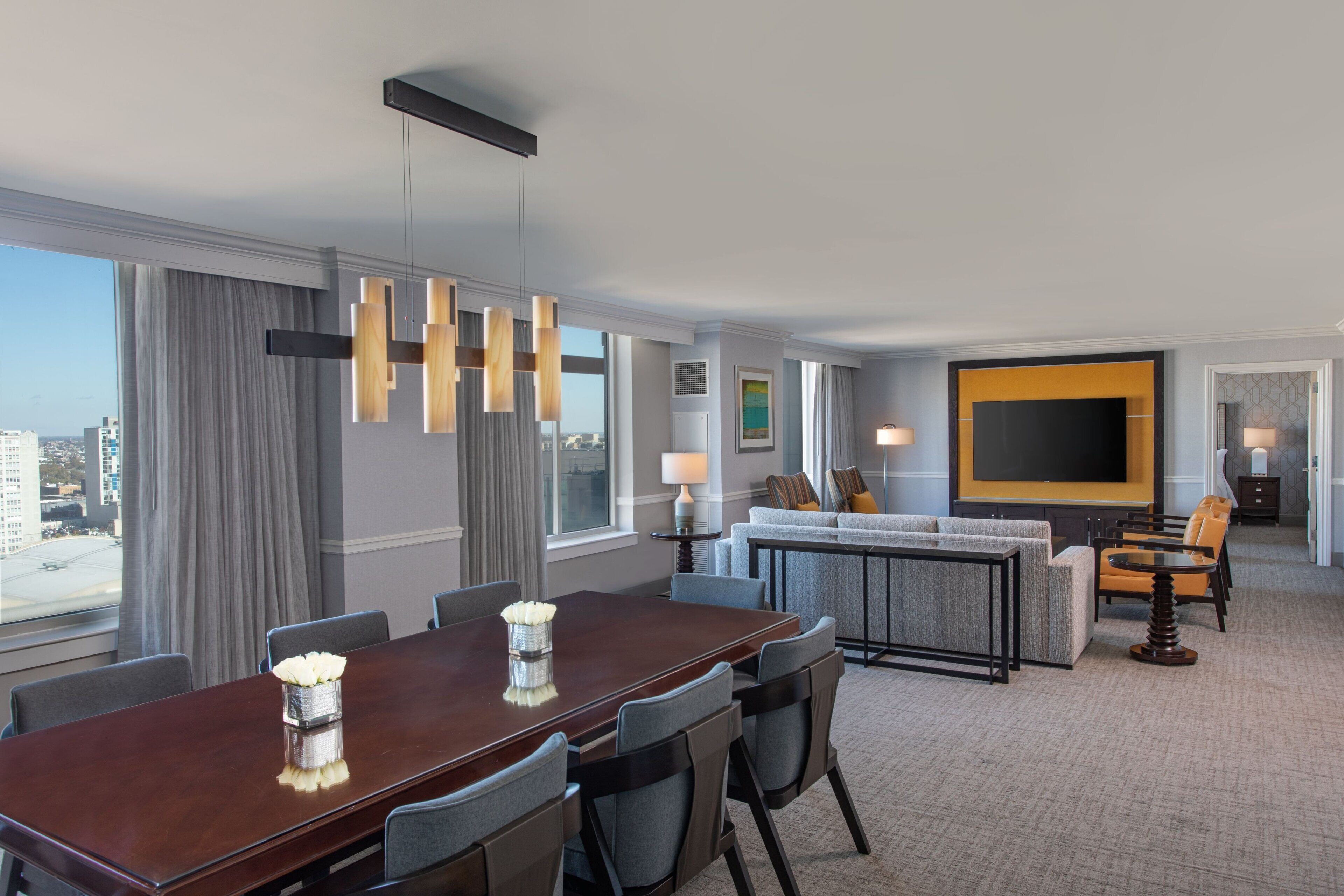 A hotel room with a dining area, a sofa set, a television, and a large window with curtains.