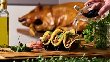 A wooden table with tacos, a bottle of oil, and a roasted pig in the background.