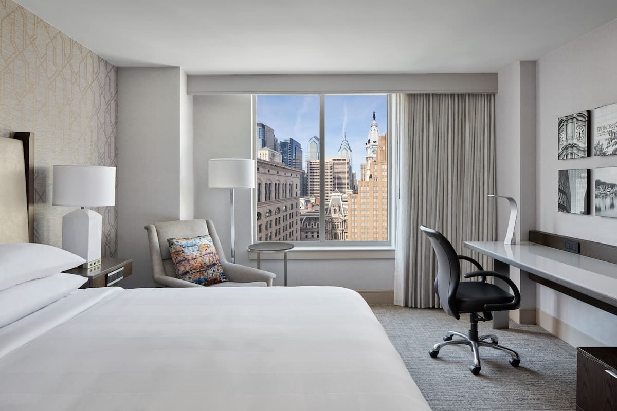 Une chambre d’hôtel avec un grand lit, un bureau, une chaise, une lampe et une vue sur le paysage urbain, visible à travers une grande fenêtre.