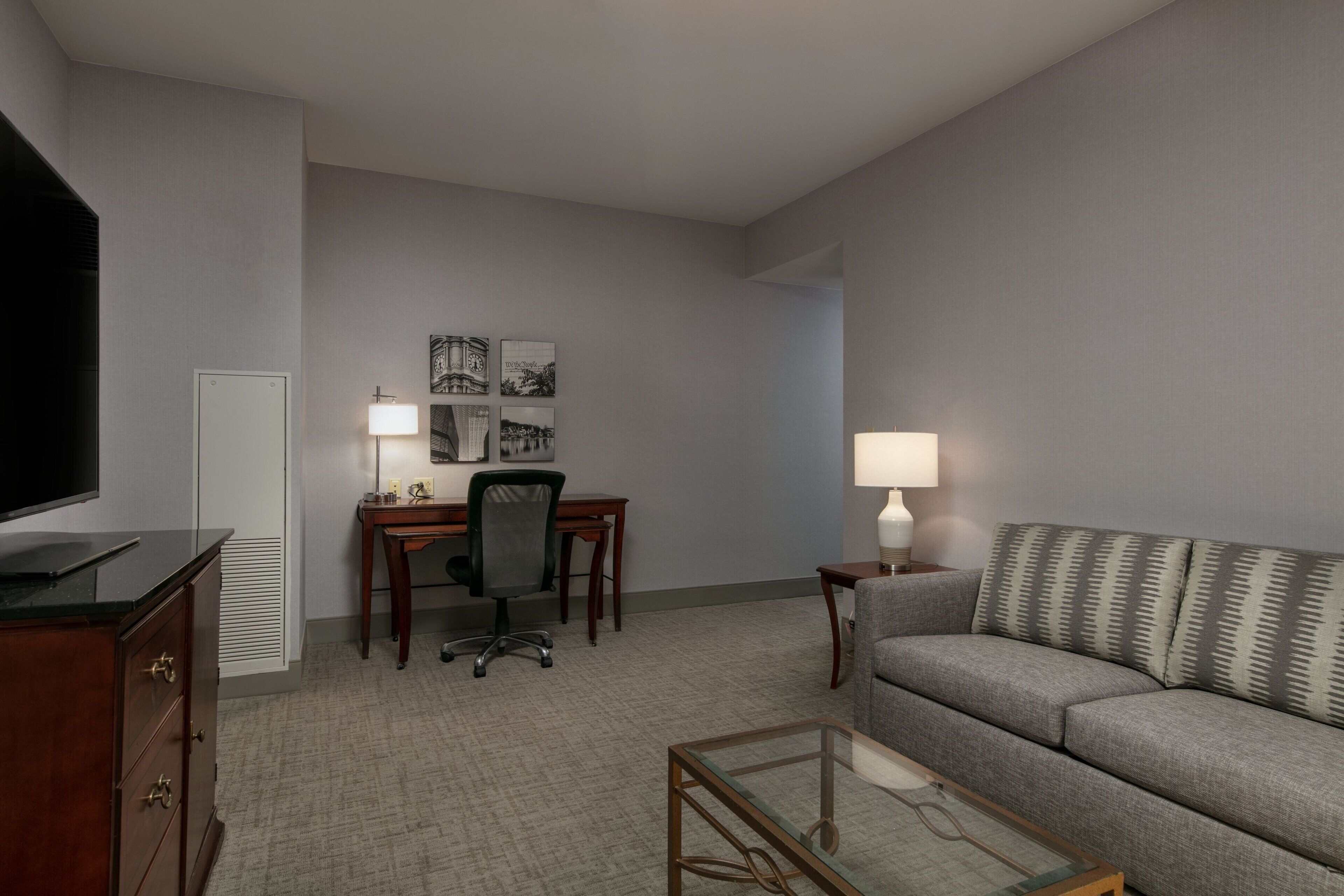 Quarto de hotel com sofá cinzento, mesa de centro de vidro, escrivaninha com cadeira, televisão e duas luminárias.
