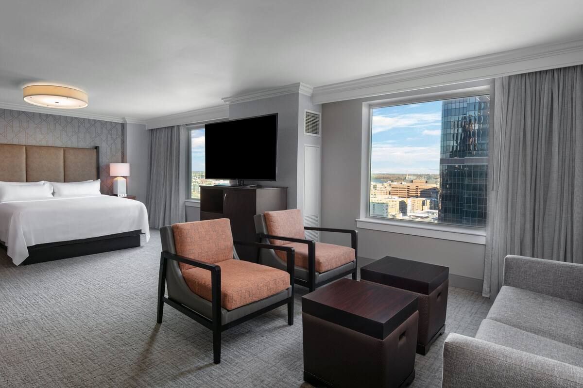 Une chambre d’hôtel comprenant un lit, deux fauteuils, une table basse, un téléviseur et une grande fenêtre donnant sur la ville.