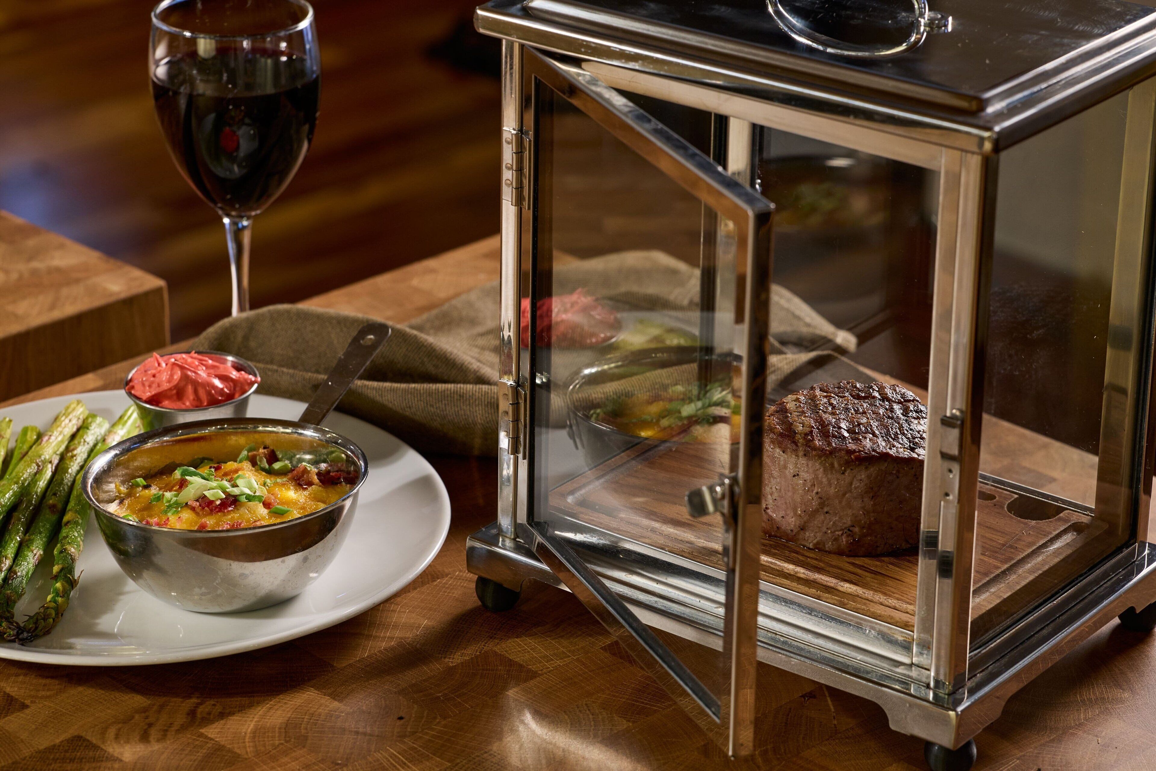 Uma mesa de jantar com uma vitrine de vidro contendo um bife, uma tigela de sopa e aspargos.