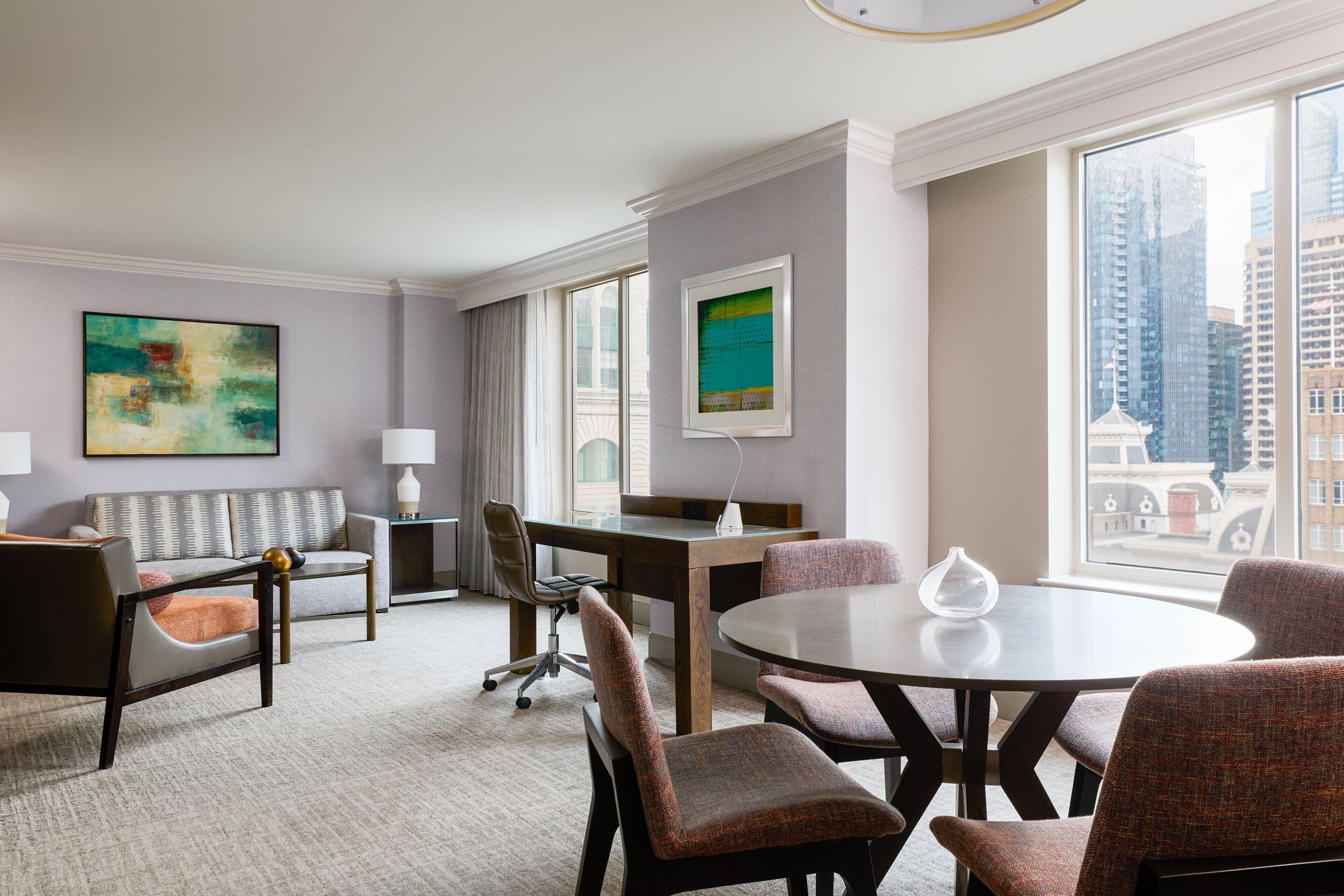 A modern hotel room with a sofa, armchairs, a dining table, and a view of the cityscape.