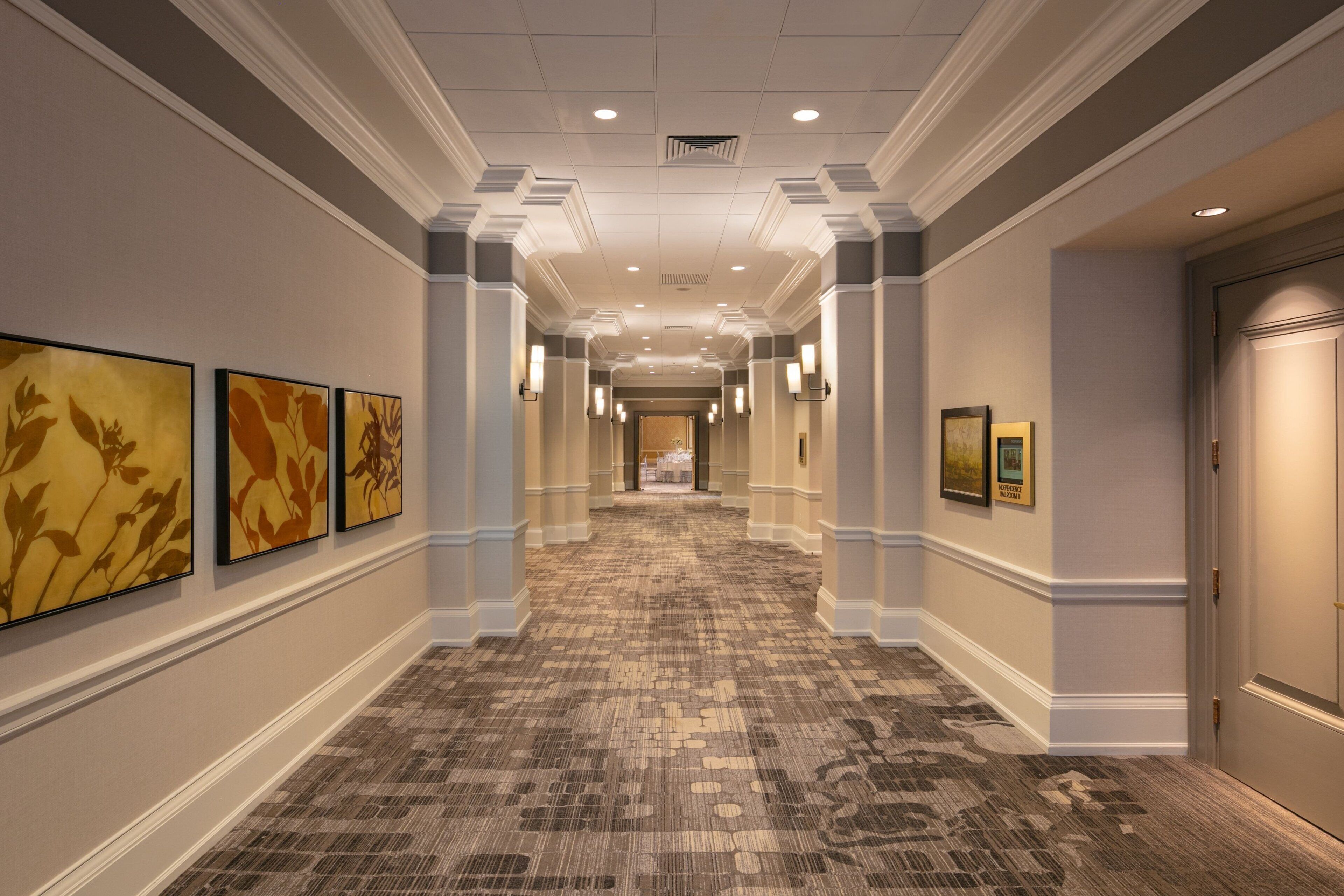 A hotel corridor with framed artwork on the walls, a patterned carpet, and a door at the end.