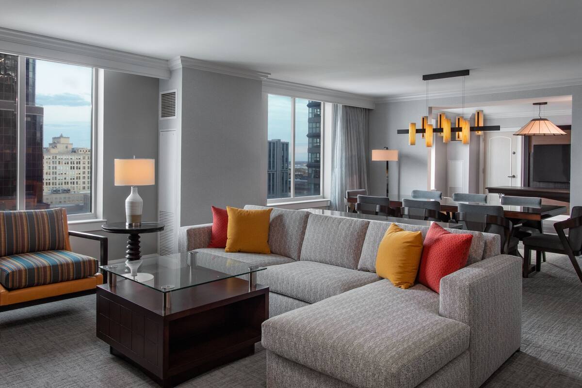 Une chambre d’hôtel moderne avec un canapé d’angle, une table basse en verre et un coin repas équipé d’un téléviseur.