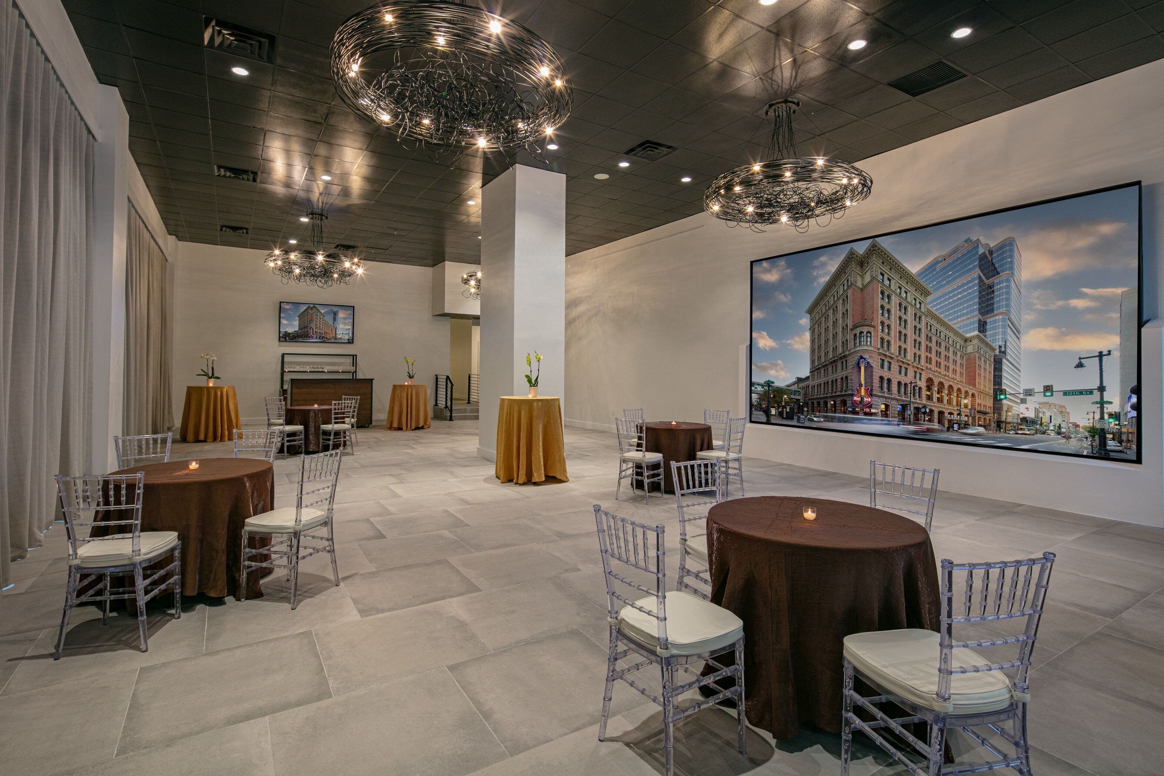 A spacious hotel lobby with multiple round tables and chairs, a large painting of a cityscape, and modern lighting fixtures.
