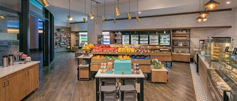 A modern cafe interior with wooden flooring, a central seating area, and a variety of food items displayed on shelves and counters.