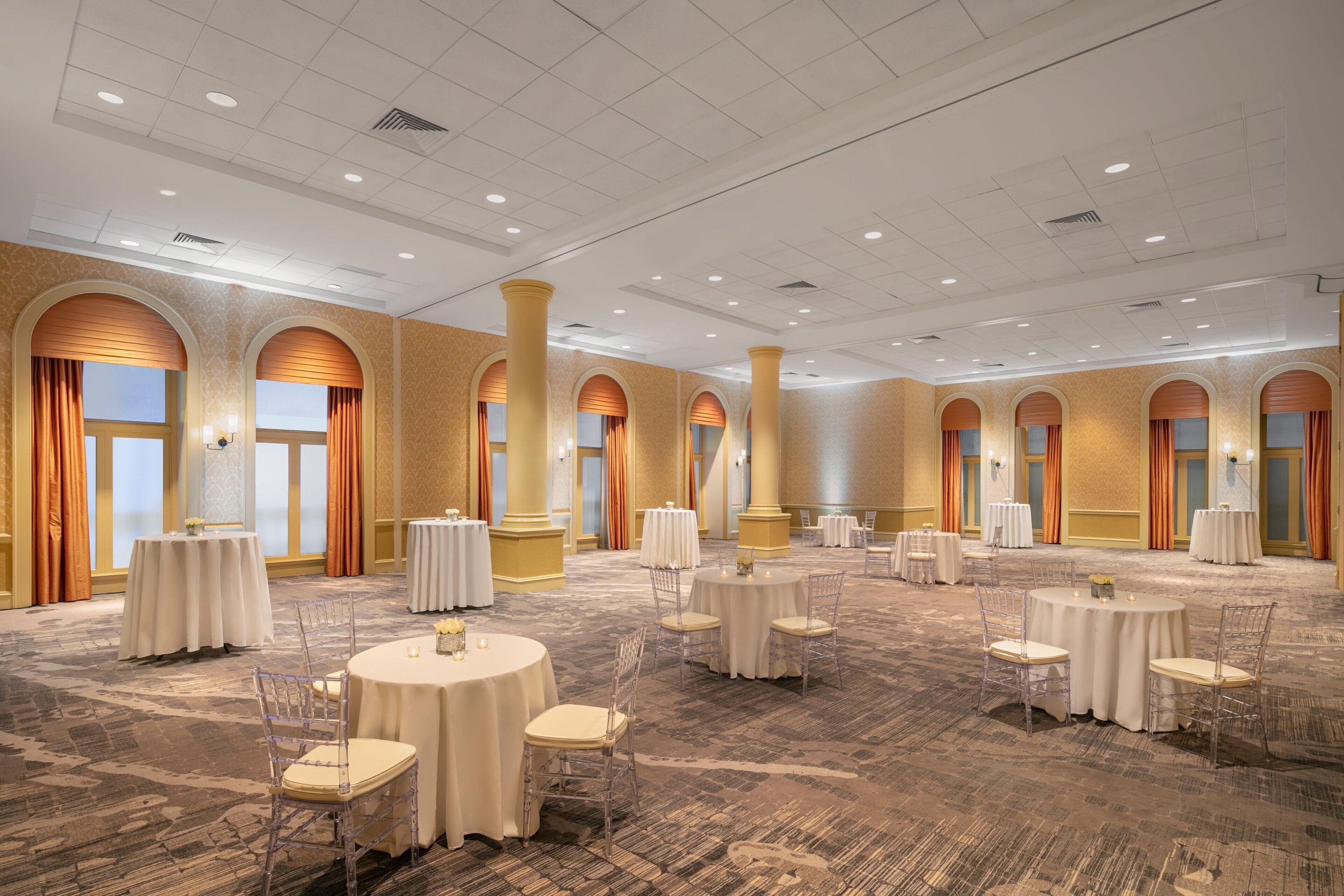 A spacious banquet hall with multiple round tables and chairs, large windows with curtains, and a patterned carpet.