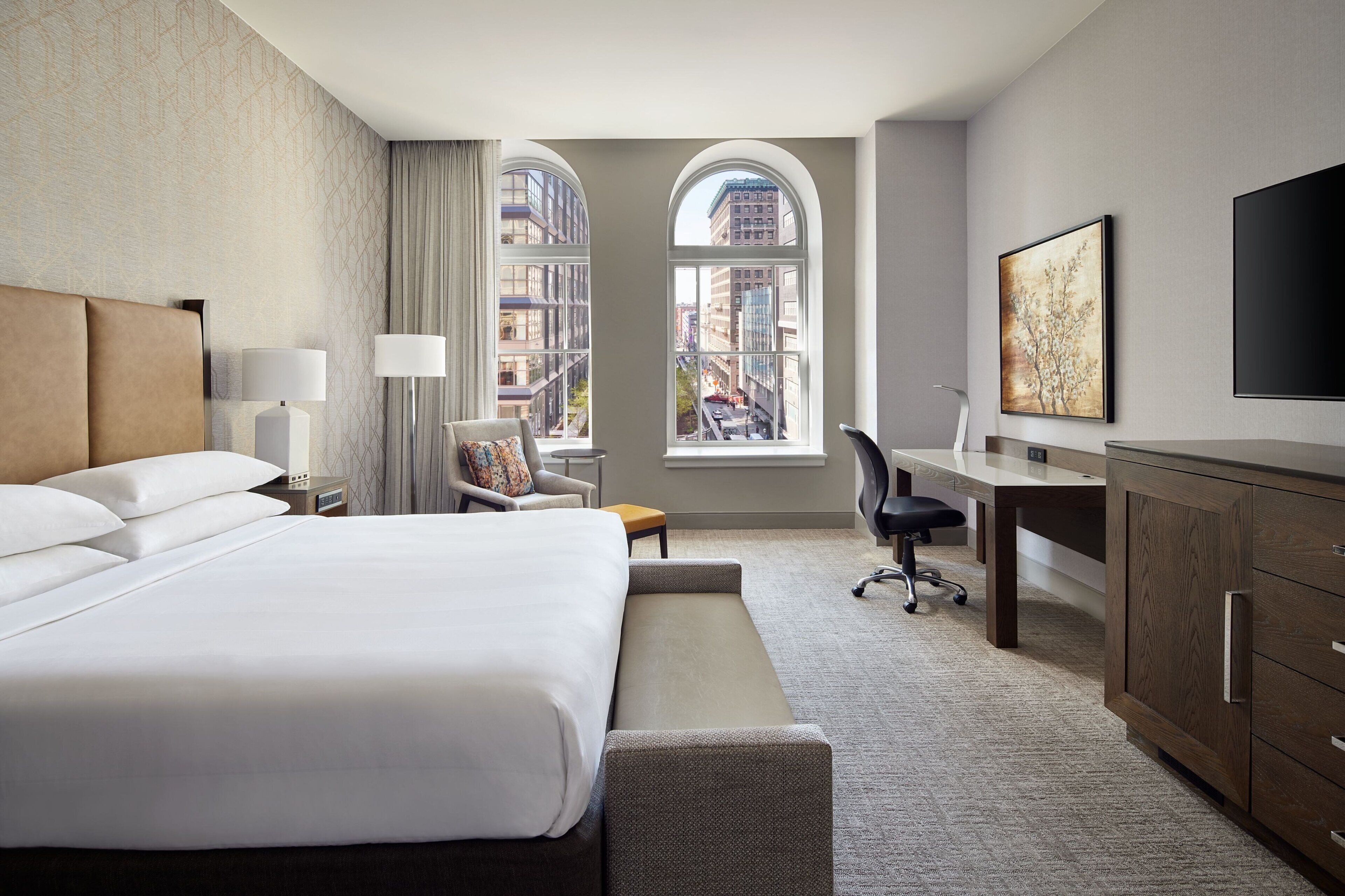 A hotel room with a large bed, a desk, a chair, a TV, and a window with a city view.
