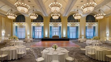 A banquet hall with round tables, chairs, chandeliers, and a stage.