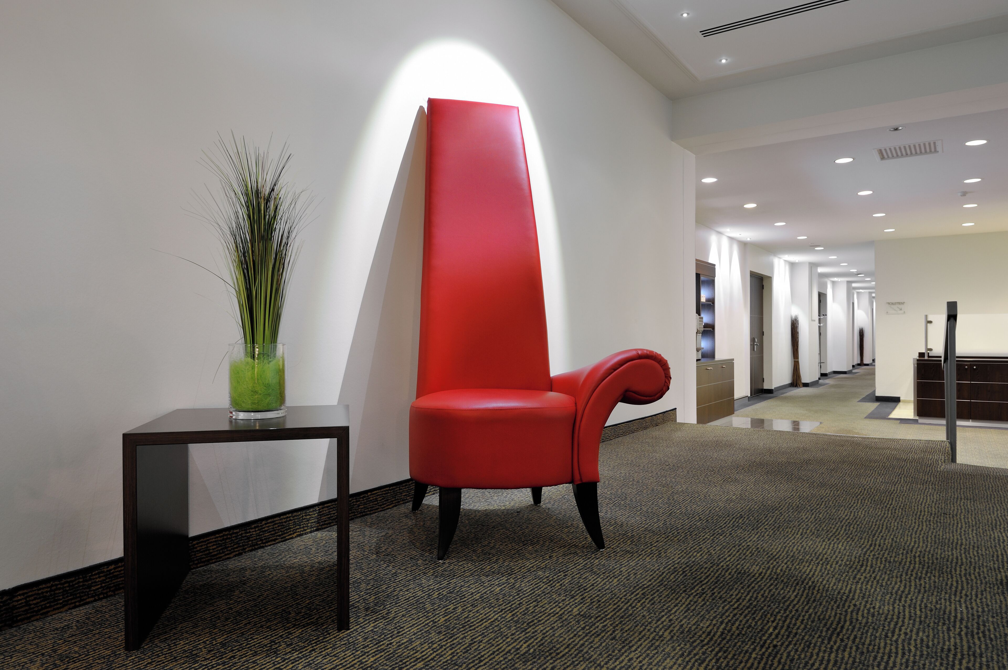 Un hall d’hôtel moderne avec un fauteuil rouge, une petite table ornée d’une plante verte et un long couloir bordé de portes.