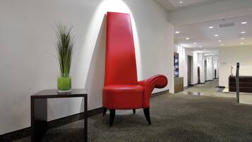 Un hall d’hôtel moderne avec un fauteuil rouge, une petite table ornée d’une plante verte et un long couloir bordé de portes.