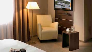 A hotel room with a bed, a chair, a TV mounted on the wall, a lamp, and a small table with a cup and glass.