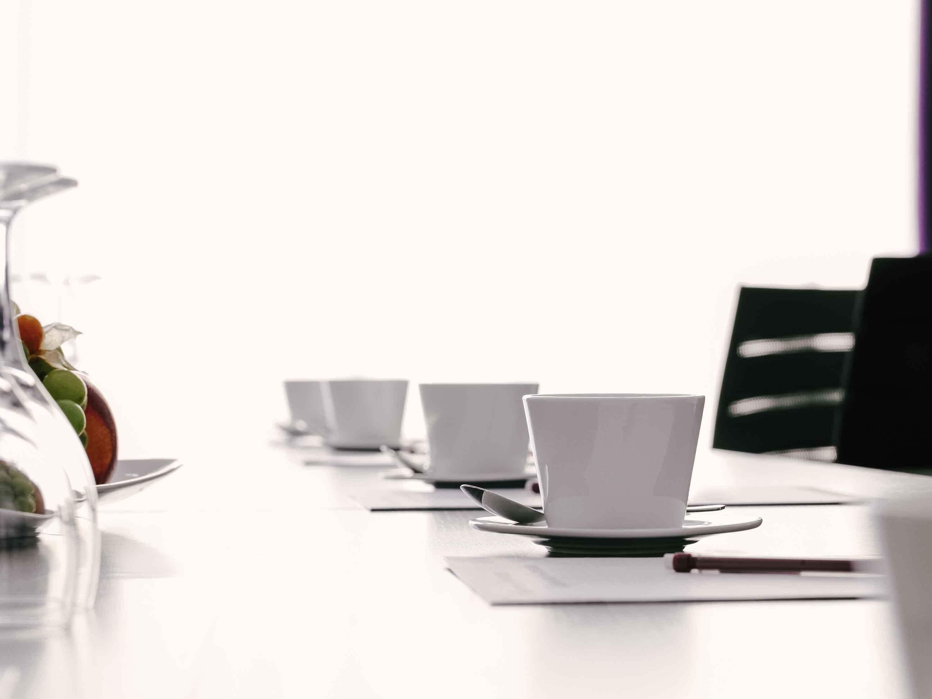 A white table with four white cups and saucers, a glass with a plant, and a notebook.