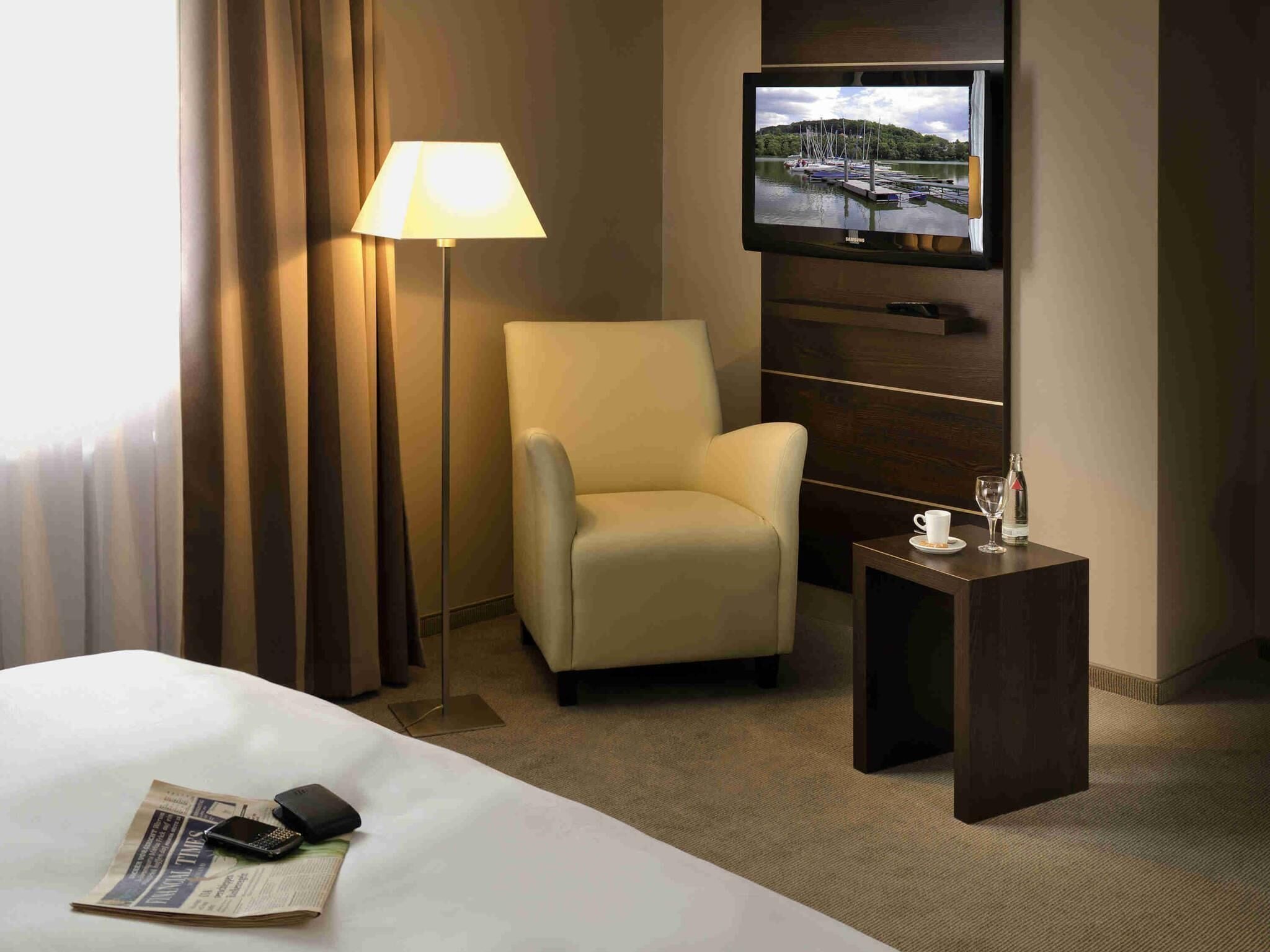A hotel room with a bed, a chair, a TV mounted on the wall, a small table with a cup and glass, and a lamp.