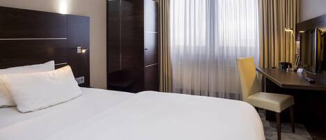 Habitación de hotel con cama, escritorio, silla, espejo y ventana con cortinas.