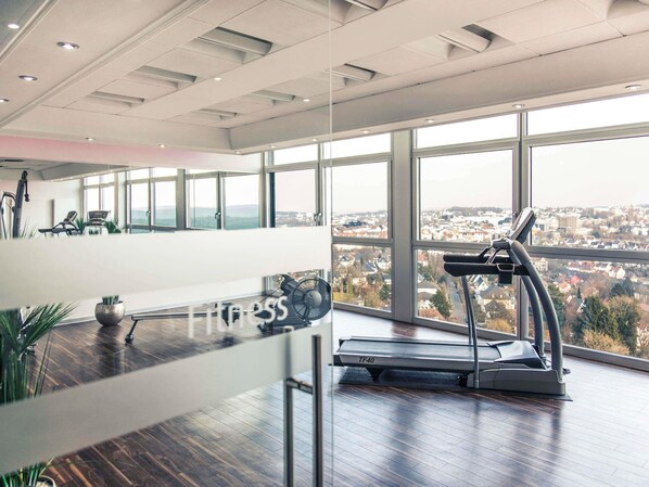 Una habitación amplia con cinta de correr, ventanas grandes y vista a un paisaje urbano.