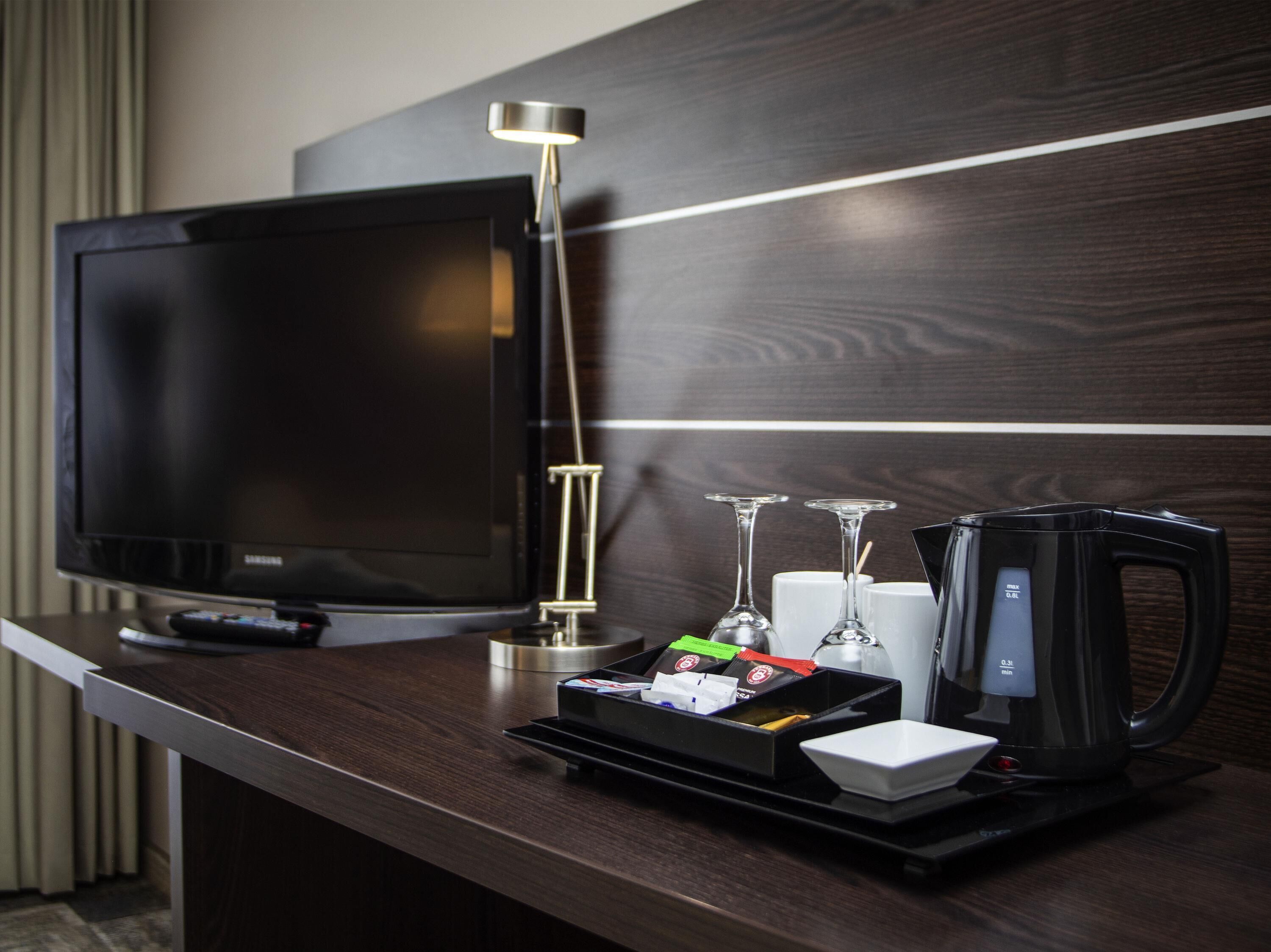 A hotel room with a flat-screen TV, a bedside lamp, a tray with a coffee maker, wine glasses, and a remote control.