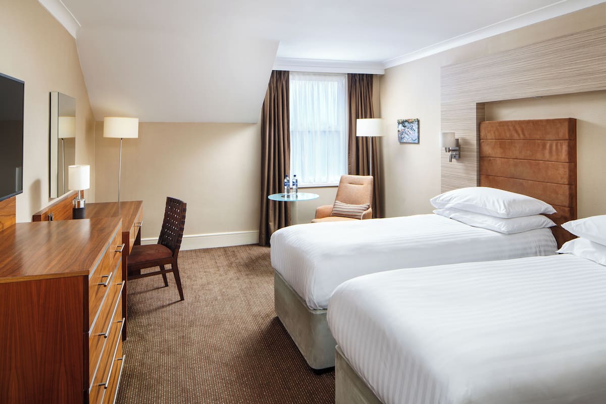 A hotel room with two beds, a desk, a chair, a small table, and a TV.