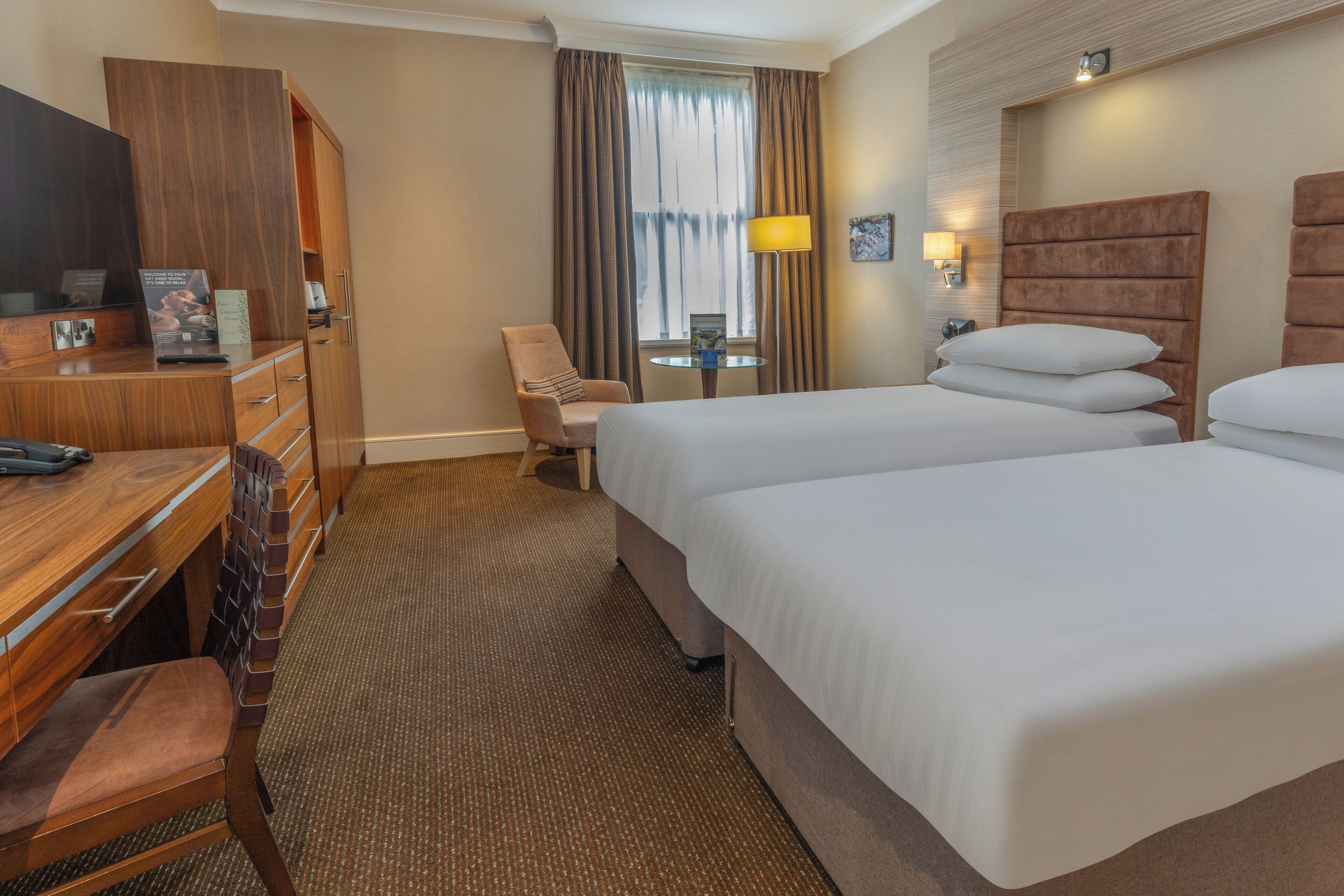 Quarto de hotel com secretária de madeira, duas camas, uma cadeira, abajur e janela com cortinas.