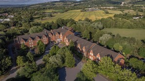 A large, red-brick building with multiple wings, surrounded by lush greenery and fields.