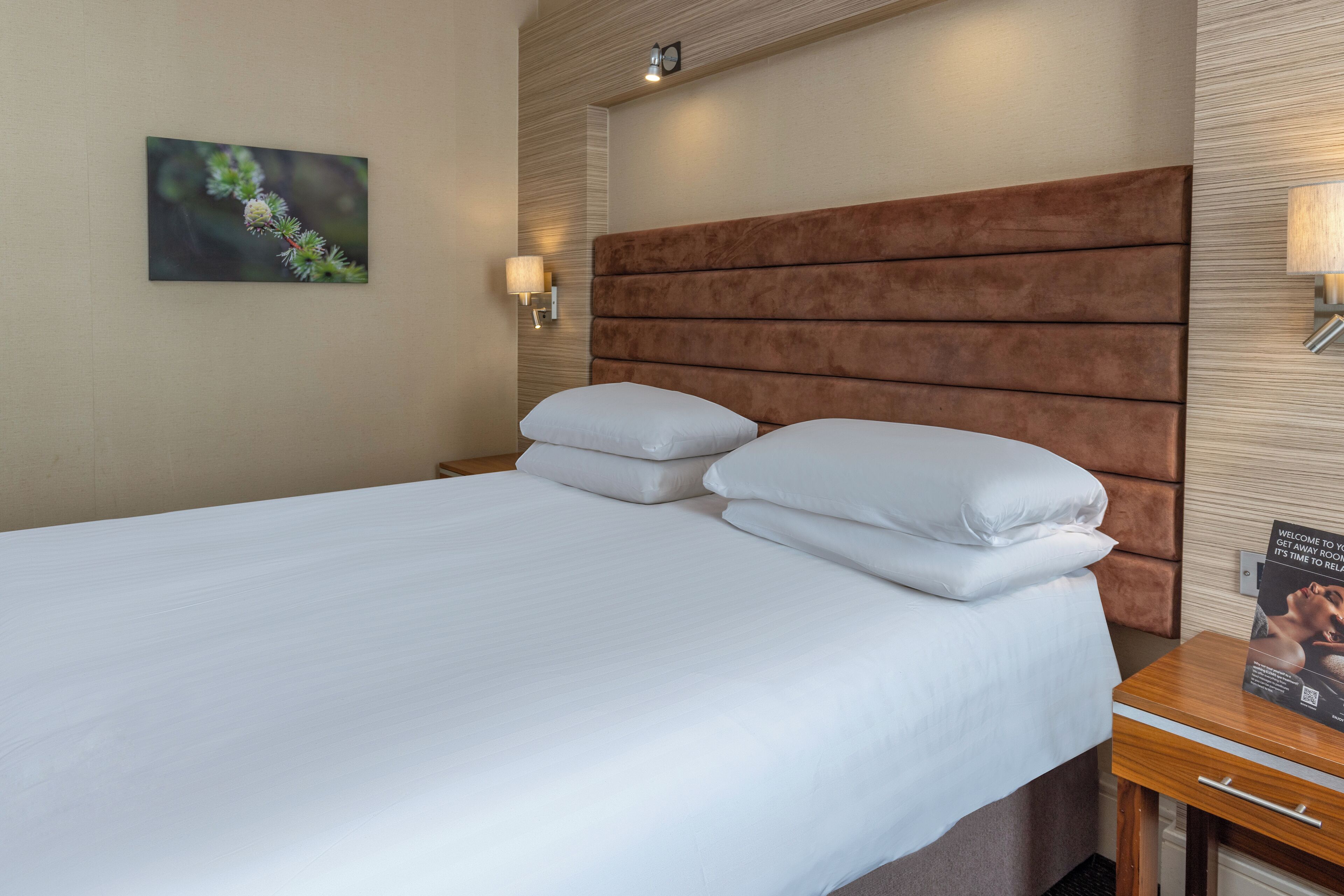 Una cama bien tendida con sábanas blancas, cabecera de madera y mesita de noche con lámpara y folleto.