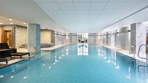 A large indoor swimming pool with clear blue water, surrounded by walls with a mix of stone and tile textures. There are lounge chairs and a small seating area with tables.