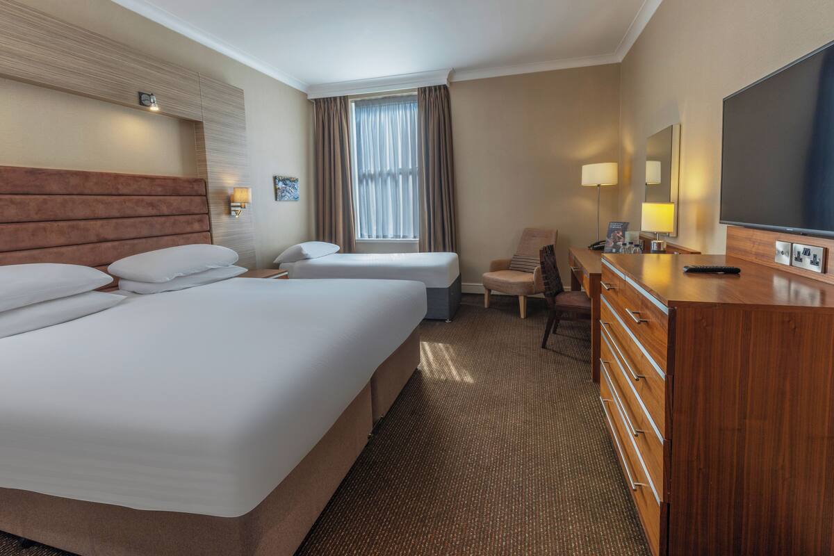 A hotel room with two beds, a desk, a chair, a dresser, and a television.