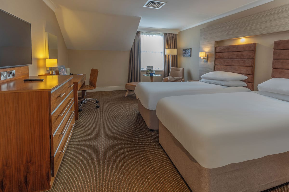 A hotel room with two beds, a desk, a chair, a television, and a window with curtains.